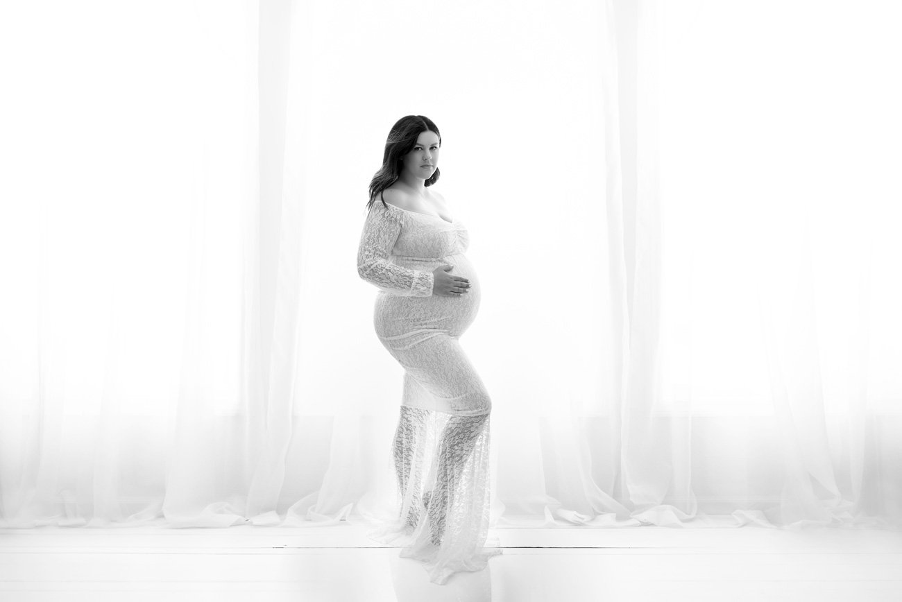 Pregnant woman standing in front of white curtain in a white lace dress